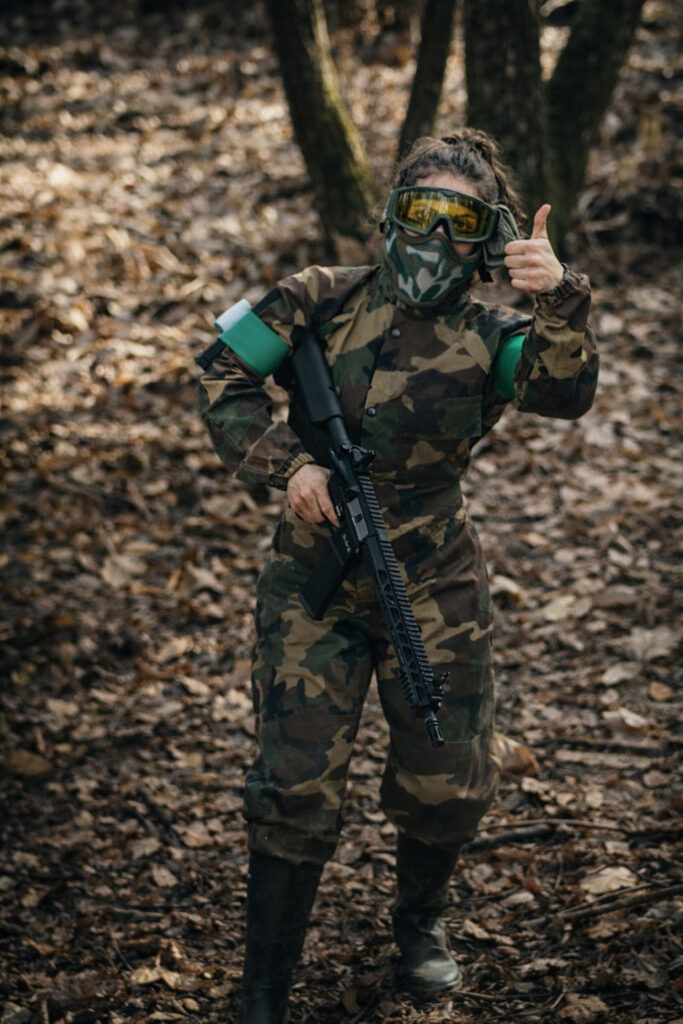 Joueuse d’airsoft en tenue camouflage faisant un signe de satisfaction lors d’une partie en forêt en Dordogne.
