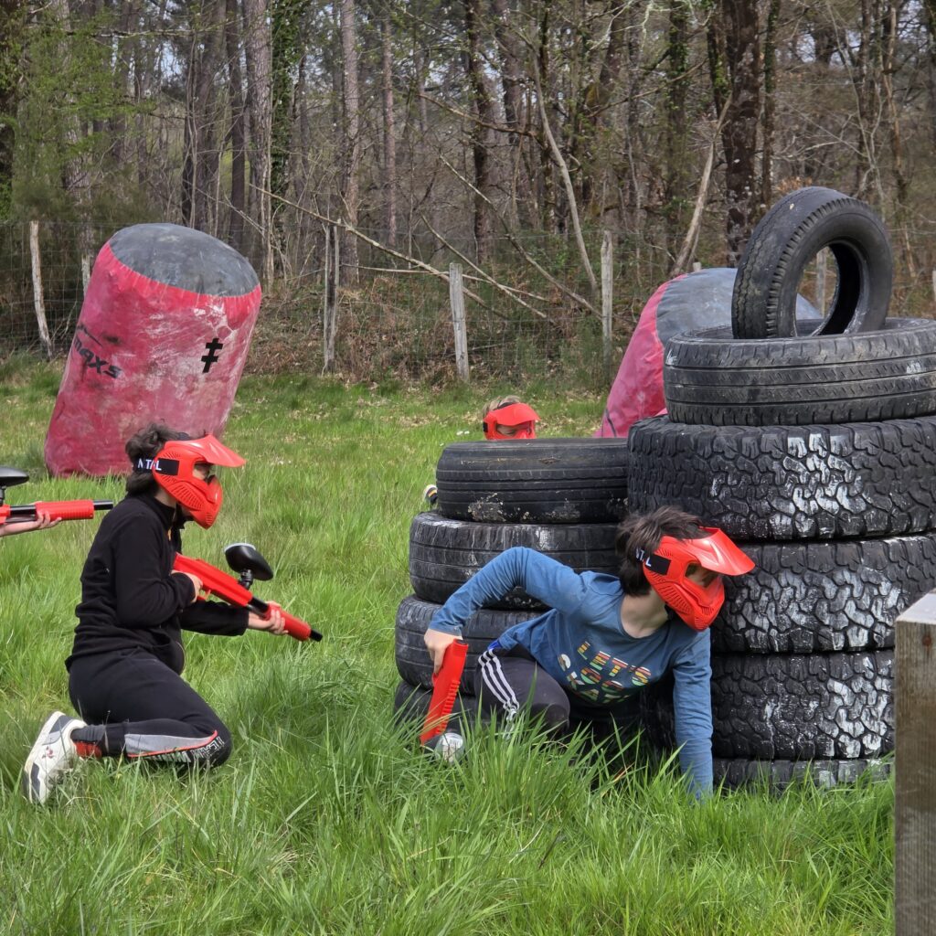 Enfants jouant au paintball derrière des obstacles en pneus et gonflables sur un terrain en Dordogne.