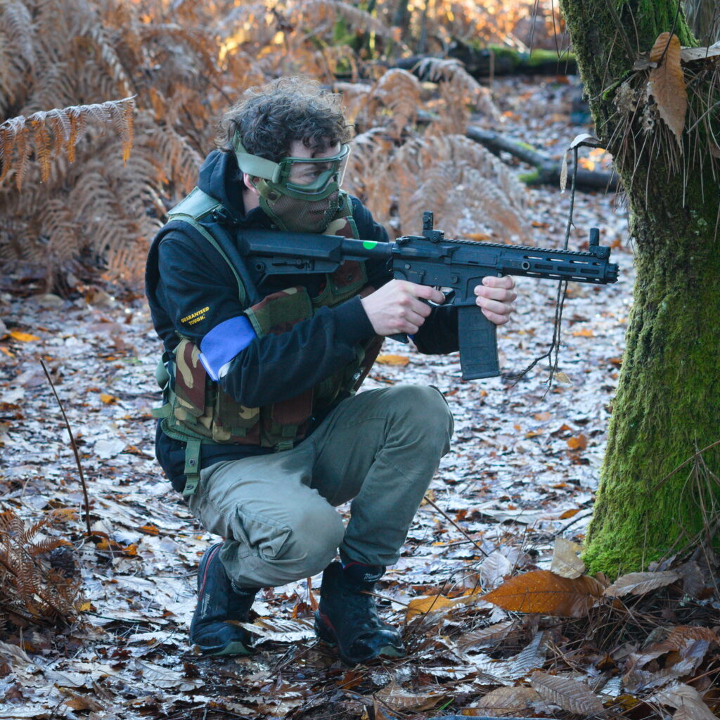 Joueur d’airsoft en position tactique derrière un arbre lors d’une partie en forêt en Dordogne.